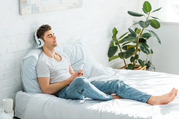 handsome man in white t-shirt and jeans sitting on bed, holding smartphone and  listening music in...