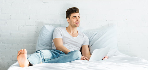 panoramic shot of handsome man in white t-shirt and jeans sitting on bed with laptop in bedroom