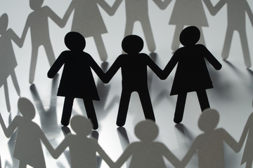 Three human paper figures surrounded by circle of paper people holding hands on white surface....