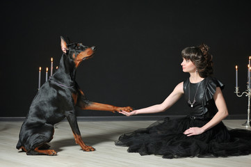 A girl in a black dress is sitting and holding a large Doberman paw against a black wall. The girl...