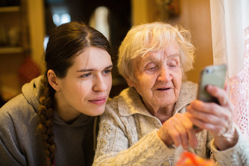Elderly woman looks and typing on a smartphone, with his adult girl granddaughter.