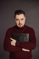 a man with a beard in casual clothes poses on an isolated dark won, in a burgundy sweater in the studio with a book