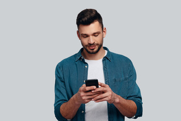 Texting to friend. Charming young man using his smart phone and smiling while standing against grey background