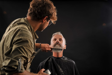 barber cuts a beard to a client to an elderly gray-haired man