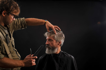 Fototapeta na wymiar Hairdresser cuts senior citizen with a beard on a dark background