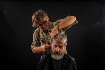 Hairdresser cuts senior citizen with a beard on a dark background