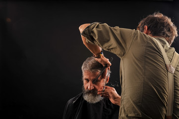 Hairdresser cuts senior citizen with a beard on a dark background