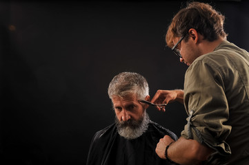 Hairdresser cuts senior citizen with a beard on a dark background