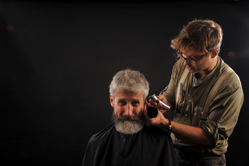 Hairdresser cuts senior citizen with a beard on a dark background in the studio