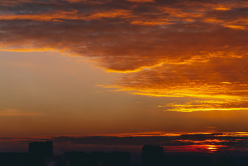 Cityscape with vivid fiery dawn. Amazing warm dramatic cloudy sky above dark silhouettes of city building roofs. Orange sunlight. Atmospheric background of sunrise in overcast weather. Copy space.