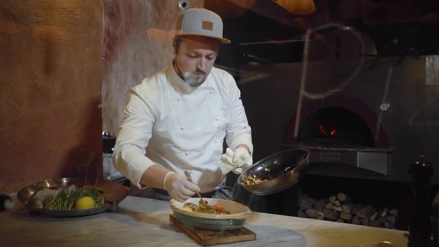 Chef in white uniform putting cutted on small pieces steamed vegetables on the cooked fish lying in deep plate in kitchen. Food preparation in modern restaurant behind glass with signature