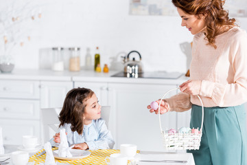 Curly woman holding wicker basket with easter eggs and looking at daughter in kitchen