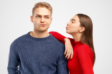 Pleased pretty female, stands sideways, leans at shoulder of her handsome bearded boyfriend, going to kiss in cheek, expresses love and truthful feeling, isolated on white wall in studio