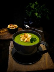 green cream of broccoli soup with wheat croutons in a black mug on a black background close-up
