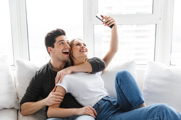 Obraz premium Portrait of positive couple man and woman taking selfie photo on smartphone while lying on couch at home