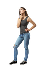 Young woman in sleeveless top and blue jeans standing in half-turn with left hand on hip and right arm rubbing under chin isolated on white background.