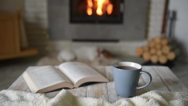 Hygge concept with open book and cup of tea near burning fireplace