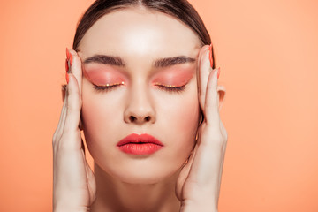 beautiful trendy young woman with glitter makeup and eyes closed touching face isolated on coral