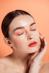 beautiful stylish young woman touching face and posing with eyes closed isolated on coral