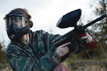 Paintball sport player girl in protective camouflage uniform and mask with marker gun outdoors