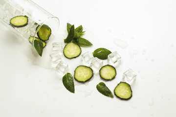 Spilled Glass with refreshing water, cucumber slices, mint leaves and ice cubes on a light background