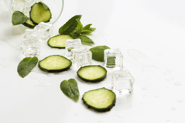 Spilled Glass with refreshing water, cucumber slices, mint leaves and ice cubes on a light background