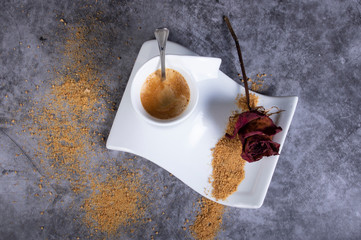 White porcelain coffee cup with brown sugar, dry rose, sugar bowl and dry rose. Hot coffee with foam.