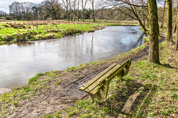 Fototapeta premium Wooden bench along a muddy trail on the bank of the river Dommel near Valkenswaard, The Netherlands on a sunny day in springtime