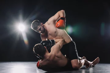 Zelfklevend Fotobehang Shirtless strong mma fighter in boxing gloves punching opponent in head while sportsman lying on floor © LIGHTFIELD STUDIOS