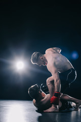 shirtless strong mma fighter in boxing gloves standing above opponent and punching him in head while sportsman lying on floor