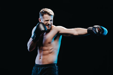 muscular sporty boxer with strenuous face expression in boxing gloves doing punch isolated on black