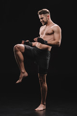 muscular barefoot mma fighter in bandages doing kick with knee on black