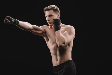 Strong shirtless muscular sportsman in bandages doing punch isolated on black