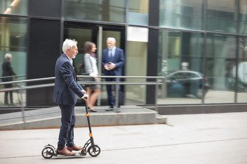 Happy senior gray-haired businessman commuting to work on a kick scooter. Riding on a sidewalk in front of an office building. Two people talking in the background, blurred