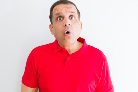 Middle age man wearing red t-shirt over white wall afraid and shocked with surprise expression, fear and excited face.