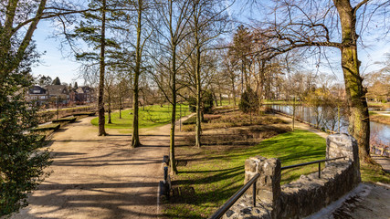 Landscape of Proosdij Park with its paths between bare trees, small river, against blue sky, view from stone bridge, sunny winter day in Meerssen, South Limburg, The Netherlands