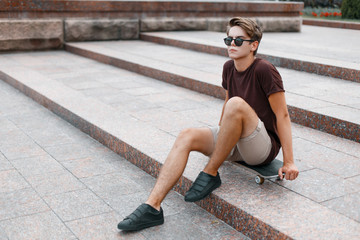 Attractive young hipster man in dark sunglasses in summer fashionable clothes in sneakers sits on a...
