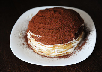 Freshly cooked tiramisu crepe cake dusted with powdery cocoa. White plate, wooden table