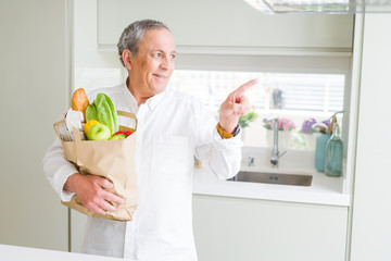 Handsome senior man holding a paper bag of fresh groceries at the kitchen very happy pointing with hand and finger to the side