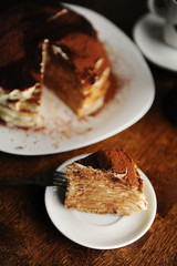 Piece of crepe cake with tiramisu filling and powdery cocoa is served on plate. The interior of coffee shop
