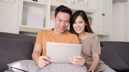 Asian couple using tablet for online shopping in internet in living room at home, sweet couple enjoy love moment while lying on the sofa when relax at home. Lifestyle couple relax at home concept.