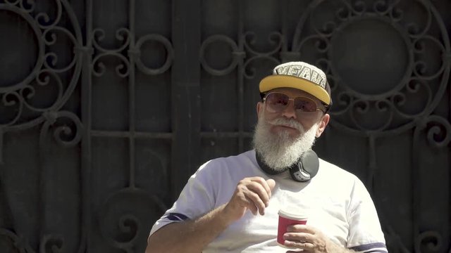 Portrait of an old bearded fashionably dressed grandfather drinking coffee and twisting his gray beard outdoors.