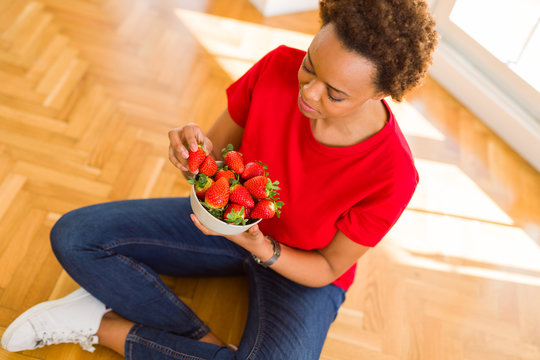 Beautiful young african woman with afro hair eating fresh strawberries sitting on the floor