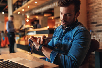 Young man sitting at cafe and working on laptop, casual businessman looking at smart watch.