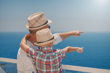 Son and dad relaxing on hotel balcony on summer holidays. They are pointing to something ahead in sea.