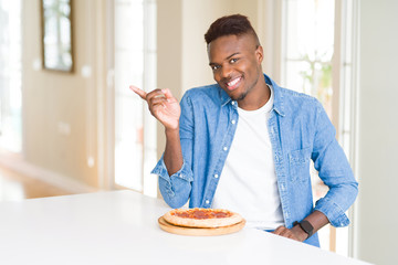 African american man eating pepperoni pizza at home very happy pointing with hand and finger to the side