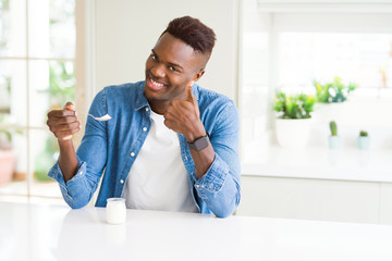 African american man eating healthy natural yogurt with a spoon happy with big smile doing ok sign, thumb up with fingers, excellent sign