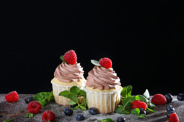 Two handmade cupcakes with blueberries and raspberries on a black background. Still life concept