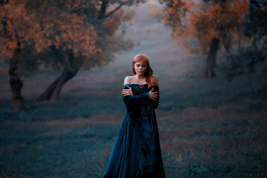 A lonely lady wanders in the fog hugging herself by the shoulders of the cold. A depressed red-haired girl in a blue dress and a gold brooch. The background nature in blue shades, yellow autumn trees