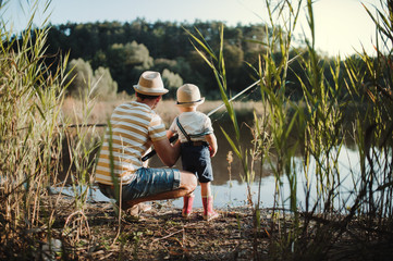 A rear view of mature father with a small toddler son outdoors fishing by a lake.